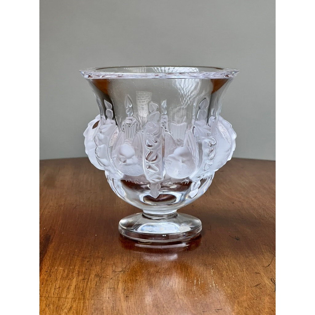A Vintage Lalique Dampierre Sparrow Footed Vase by Lalique, with a wide flared rim and intricate raised leaf and sparrow designs, stands on a wooden surface against a plain gray background.