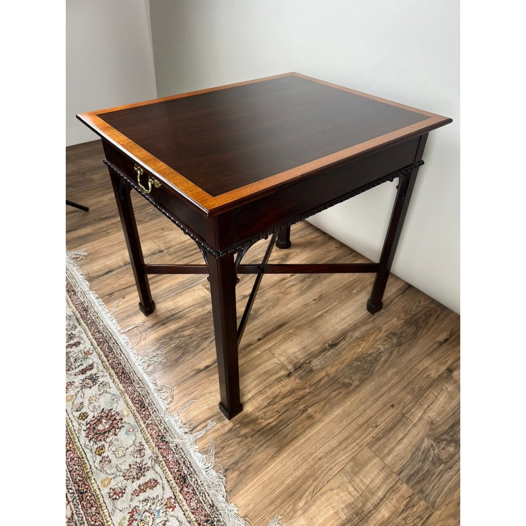 The Vintage Federal Style X Stretcher One Drawer End Table by Stickley features a dark wood finish, lighter trim, brass handle, and decorative crossbars, standing on a wooden floor next to a patterned rug.
