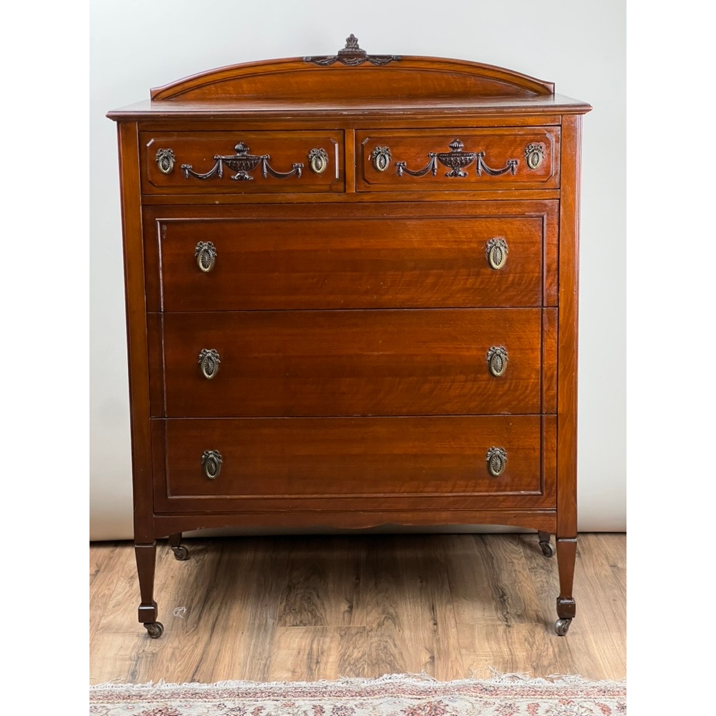 An Antique Depression Era Mahogany Two Over Three Chest of Drawers c. 1930s, featuring ornate brass handles and decorative carvings, stands on a wooden floor against a plain white wall—a classic piece of American furniture.