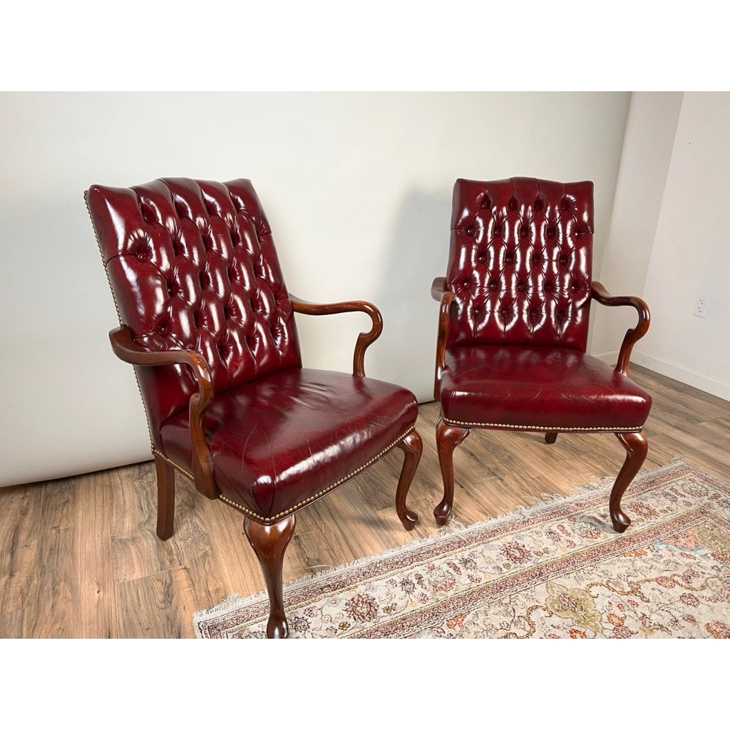 A pair of Chippendale style tufted burgundy leather library armchairs are arranged on a hardwood floor beside a patterned area rug, set against a light-colored wall.