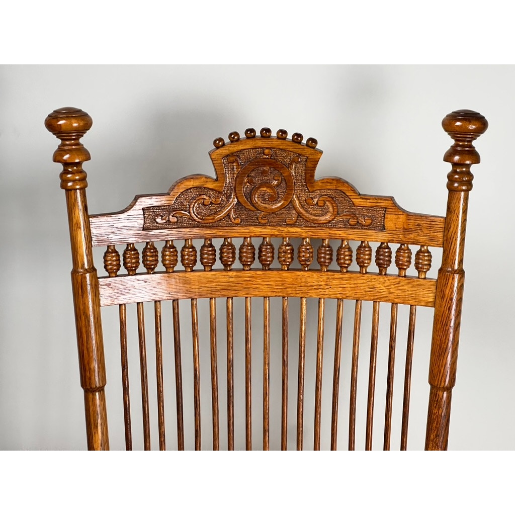Close-up of the Antique Victorian Oak Platform Rocker c. Late 19th Century, showing its ornate carved back, turned spindles, and side finials against a plain light background. Brand: None/Unknown.