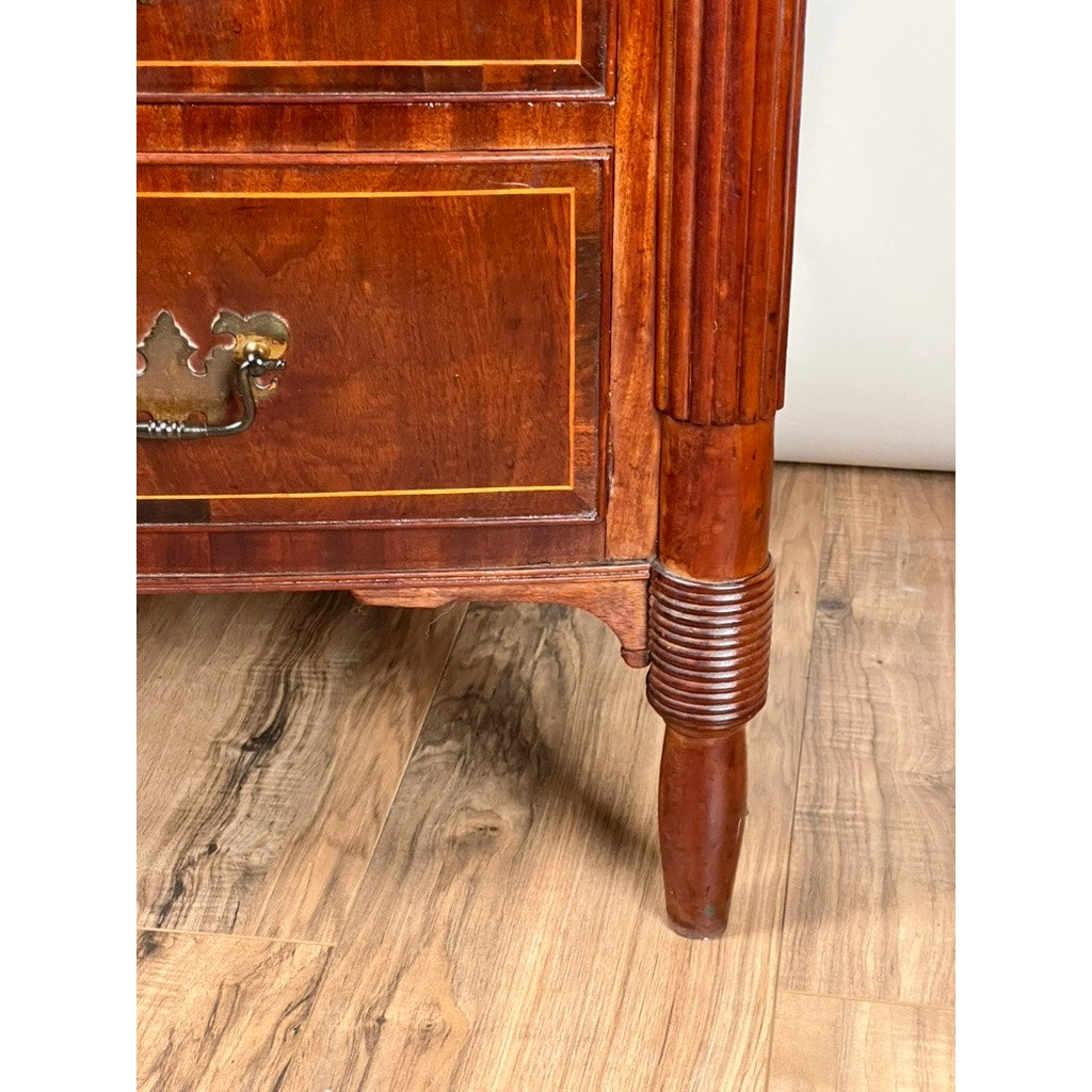 Close-up of an Antique Sheraton Mahogany Bowfront Chest c. Early 19th Century, showing a brass handle, intricate woodwork, and a tapered leg on a light floor. The polished finish highlights the grain and decorative carvings. Brand: Unknown.
