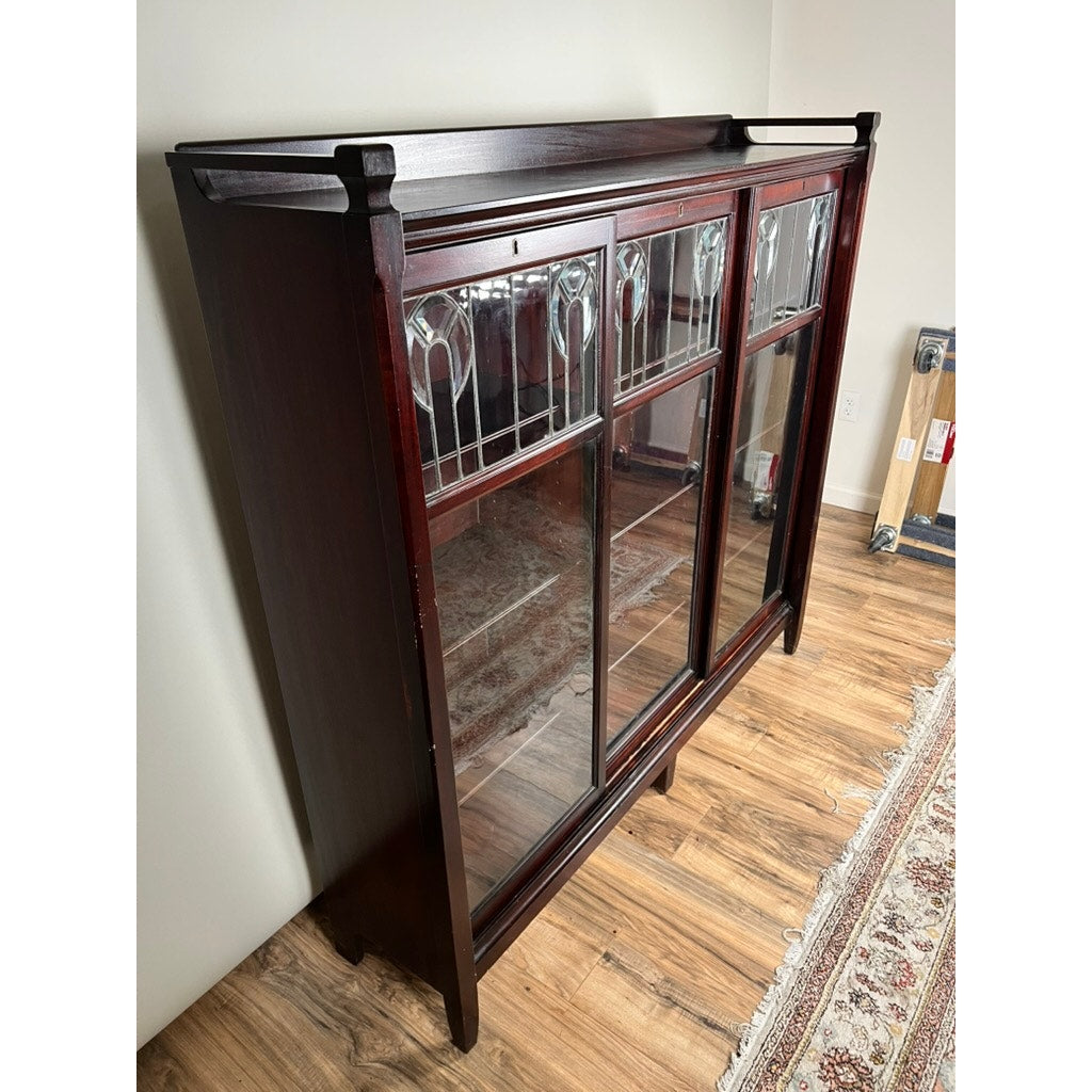 An Antique Arts & Crafts Leaded Glass Mahogany Bookcase c. 1920 stands on a hardwood floor beside a patterned rug, featuring leaded and stained glass panels at the top.