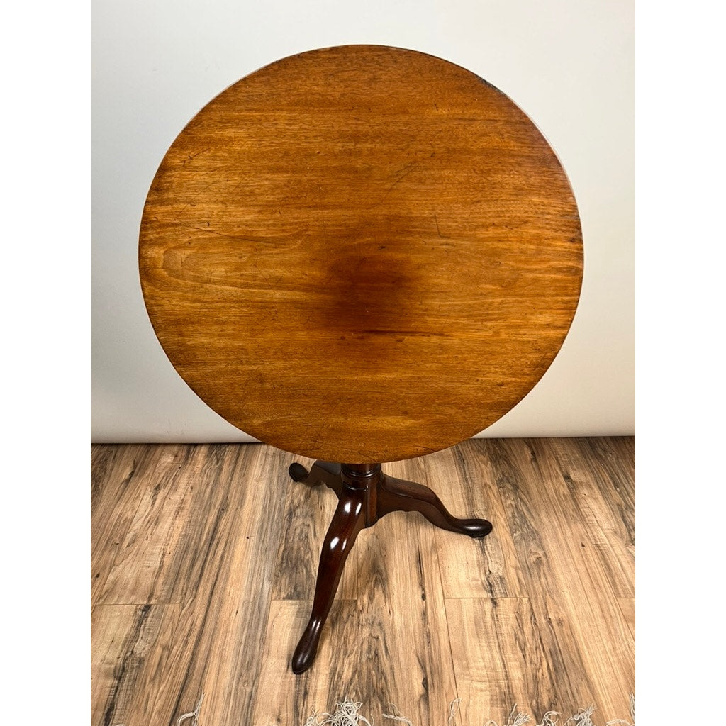 An Antique Queen Anne Mahogany Birdcage Tilt Top Breakfast Table c. Early 19th Century, with a polished surface and three curved legs, stands on a wood-patterned floor against a plain light-colored wall.