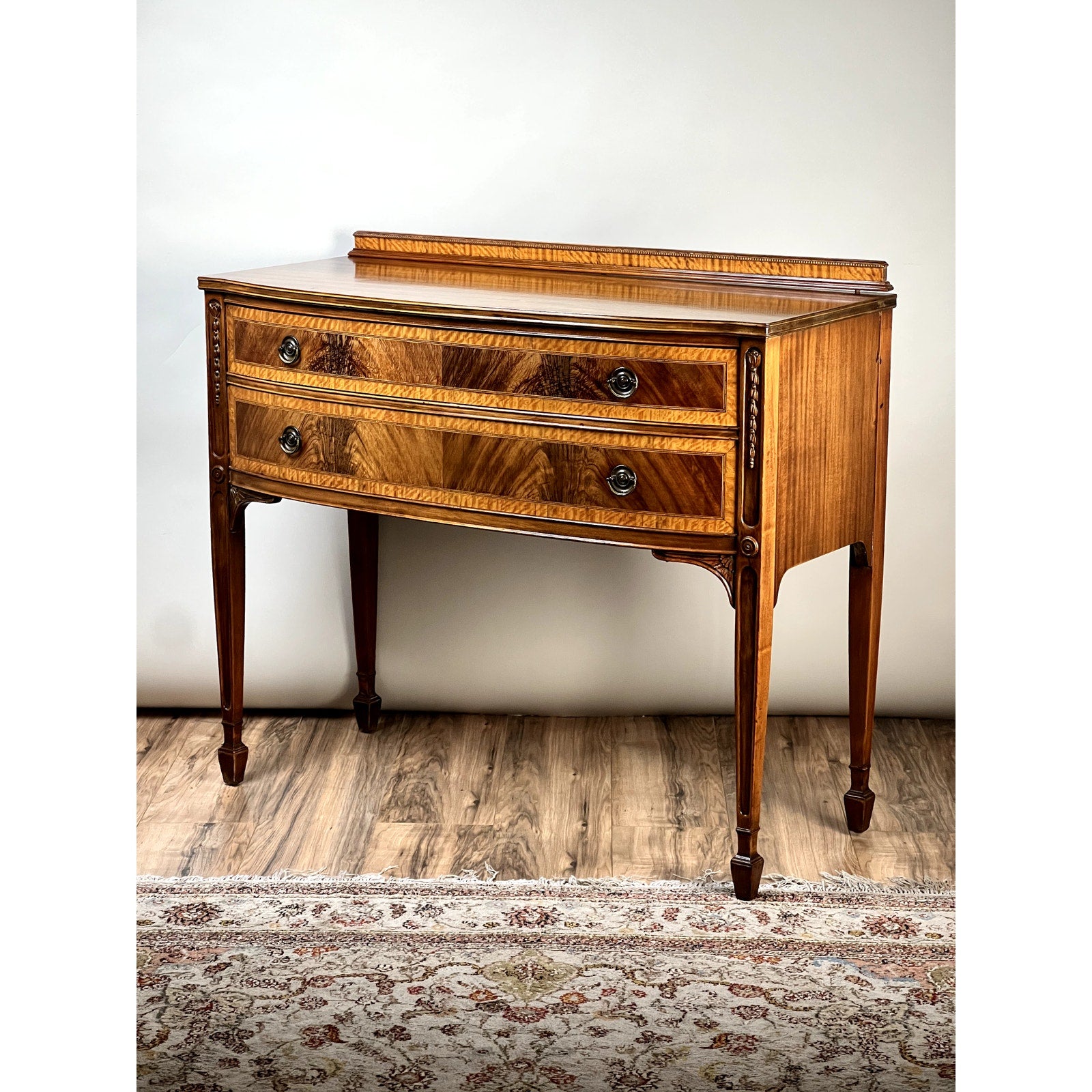 A Vintage Federal Style Bow Front Mahogany Sideboard by Paine Furniture Boston features two drawers with ornate handles and decorative inlays. It stands on tapered legs above a patterned rug and wood floor, set against a plain light background.