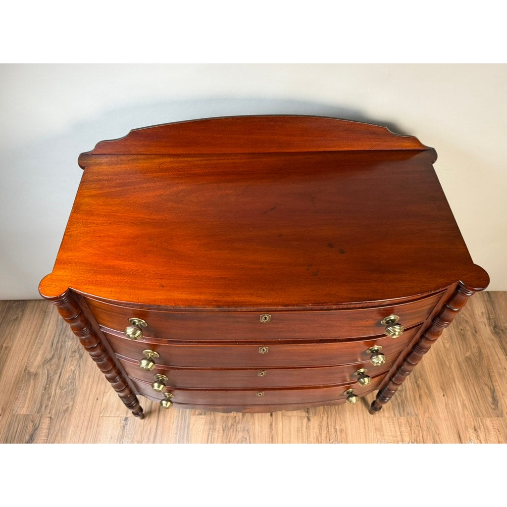 An Early 19th Century New England Federal mahogany bow-front chest of drawers with four drawers and backsplash, dating from circa 1820–1840, featuring turned side columns and brass handles, stands on a hardwood floor against a plain light-colored wall.