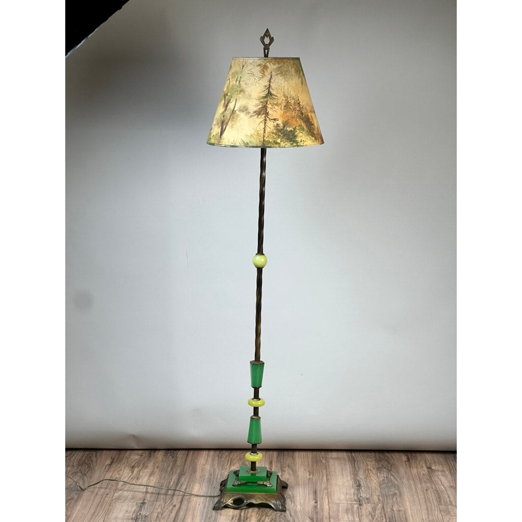 An Antique Art Deco Uranium and Slag Glass Lamp c. 1920 with a painted nature scene shade, twisted metal pole with green and yellow accents, and a decorative green and brown base stands on a wood floor against a plain backdrop.