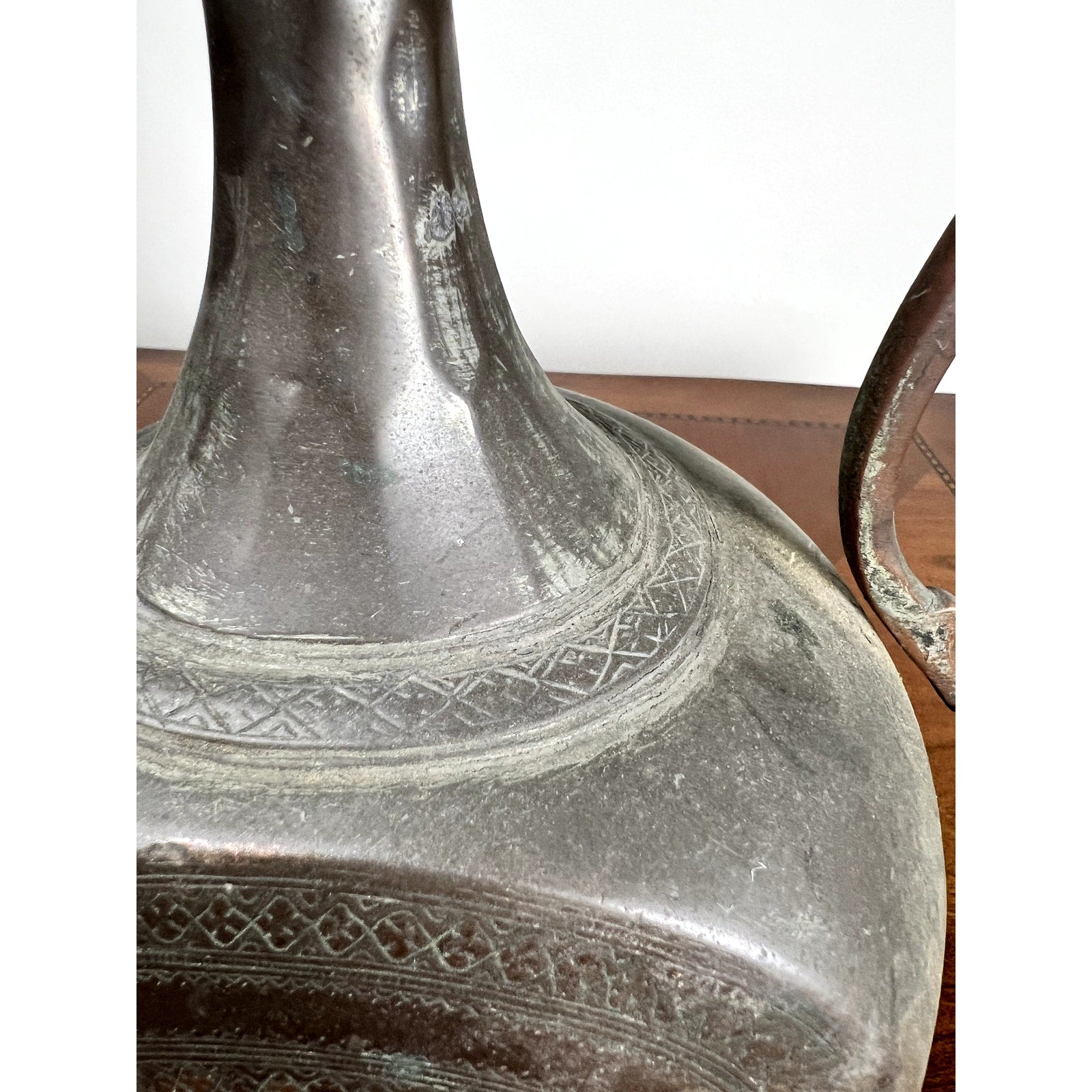 Close-up of an Antique Qajar Persian Copper Engraved Ewer (19th Century) featuring geometric patterns on its neck and body, set on a wooden surface. The handle appears partially on the right, highlighting fine Persian craftsmanship.