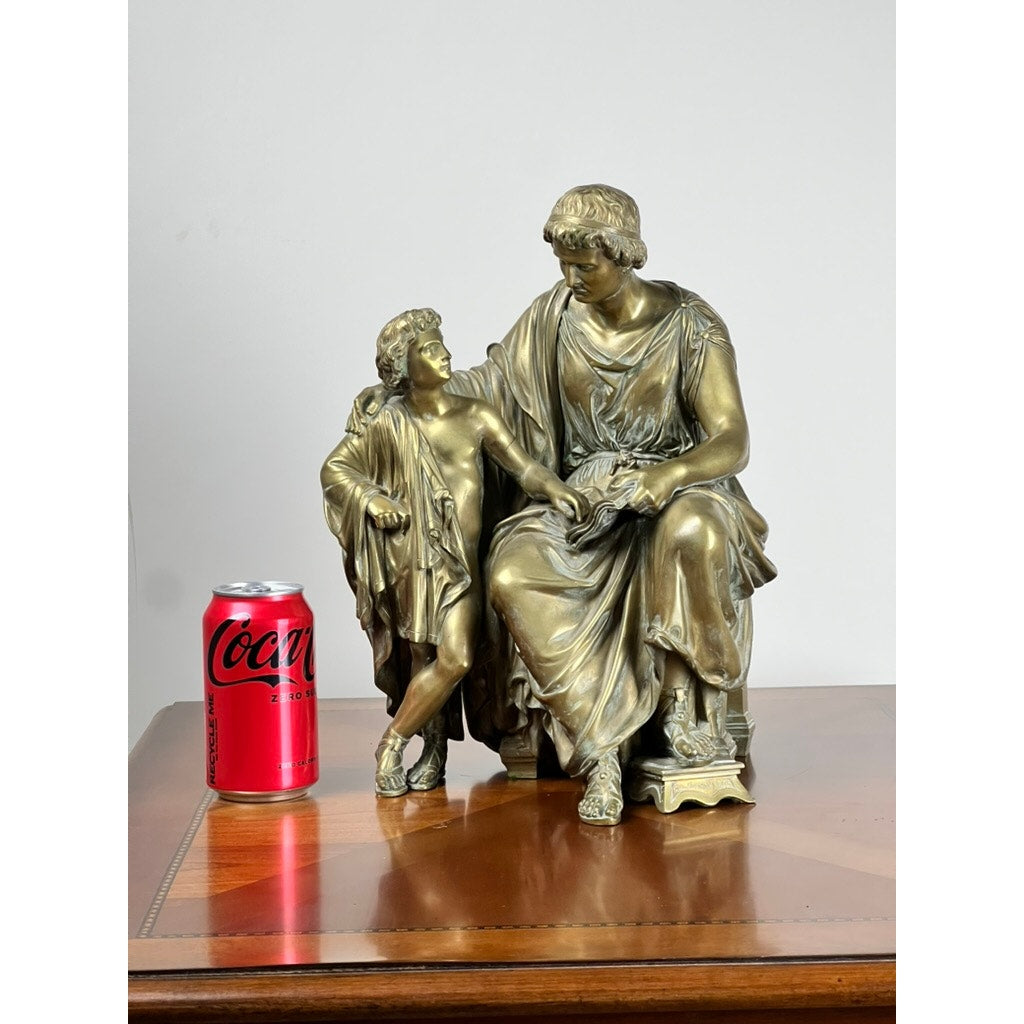 An antique bronze sculpture, “Teacher and Pupil” by Louis Charles Janson (signed Ch. Janson, 1865), depicts two figures in classical attire, seated on a wooden surface beside a red Coca-Cola can for scale.