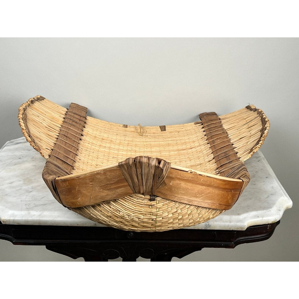 The Vintage Traditional Winnowing Basket, made of light and dark natural fibers with a curved base and wooden end supports, adds rustic farmhouse decor charm—shown here on a white marble tabletop. Brand: None/Unknown.