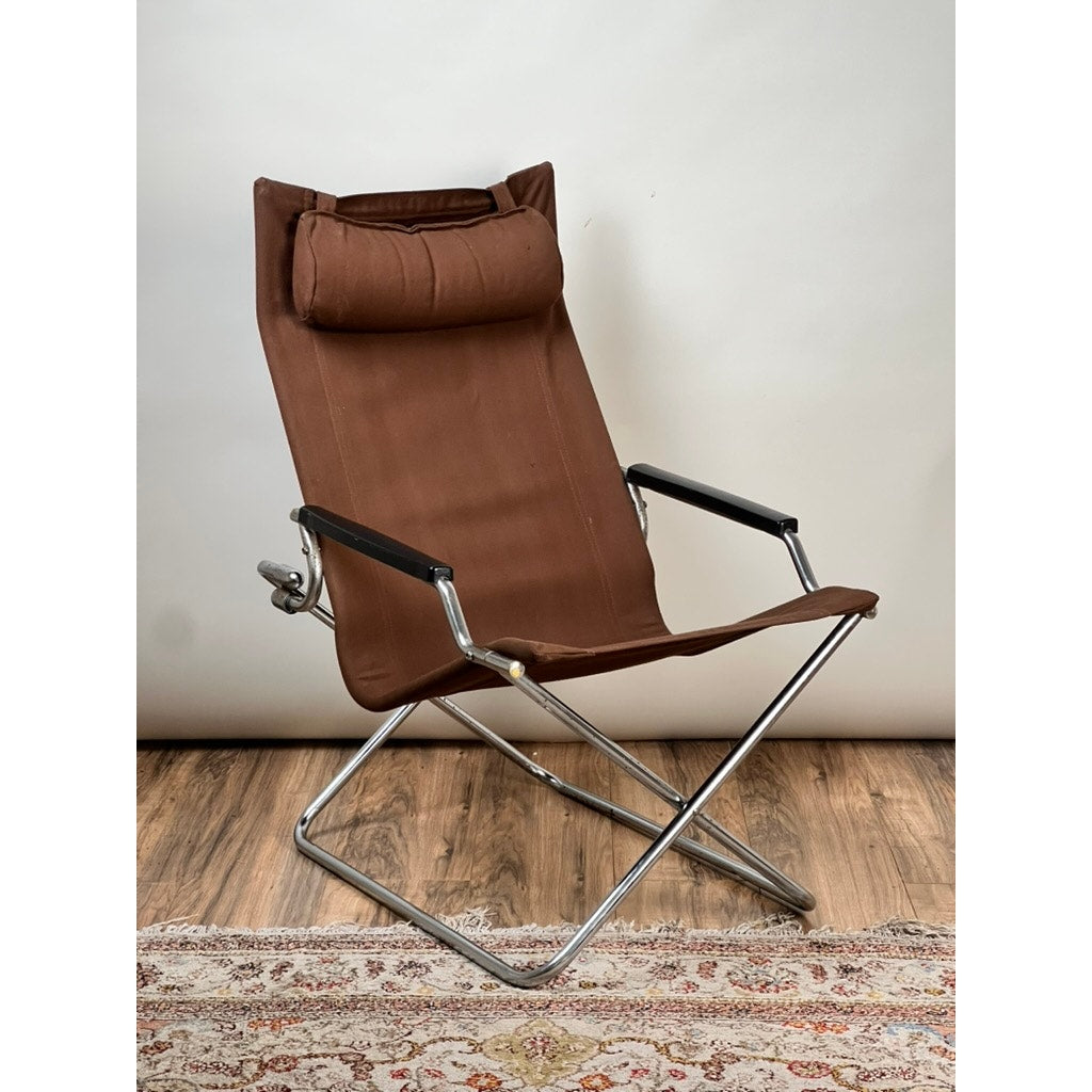 The Vintage Uchida Z Chair Japanese Modern Chrome Sling Lounge Chair with Headrest by Uchida features a brown seat, black armrests, and a sleek chrome metal frame. The vintage chair is placed on a wooden floor, with part of a patterned rug visible at the bottom, set against a plain white backdrop.