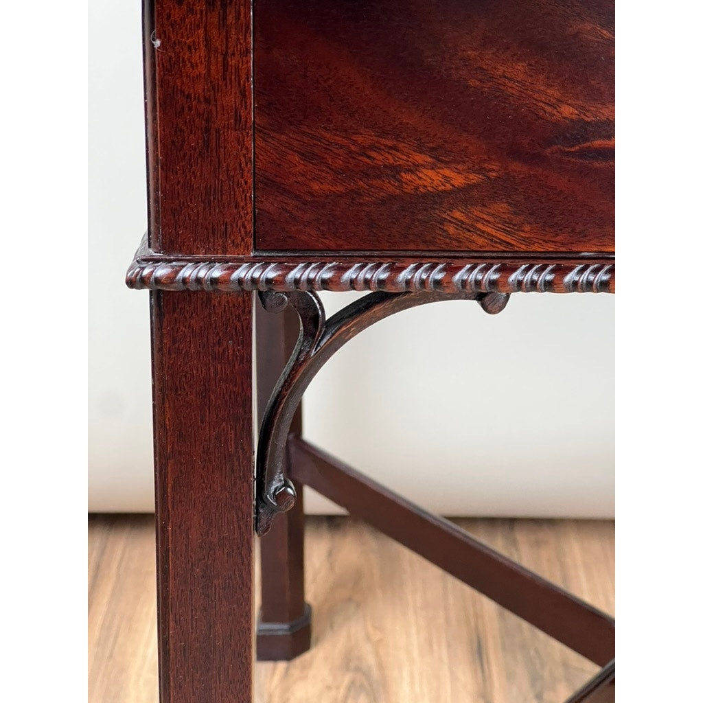 Close-up of a Stickley Vintage Federal Style X Stretcher One Drawer End Table’s antique wooden leg with carved detailing and ornate support brackets, set on a wooden floor against a neutral background.