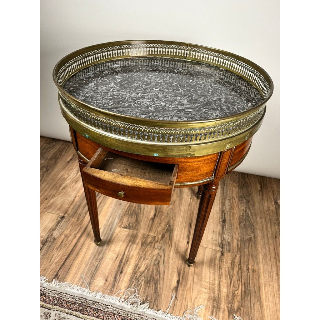 An Antique 19th Century French Louis XVI-Style Mahogany Bouillotte Table with a marble top and pierced brass gallery, featuring slender legs and a partially open drawer, stands on a wood floor beside a rug.
