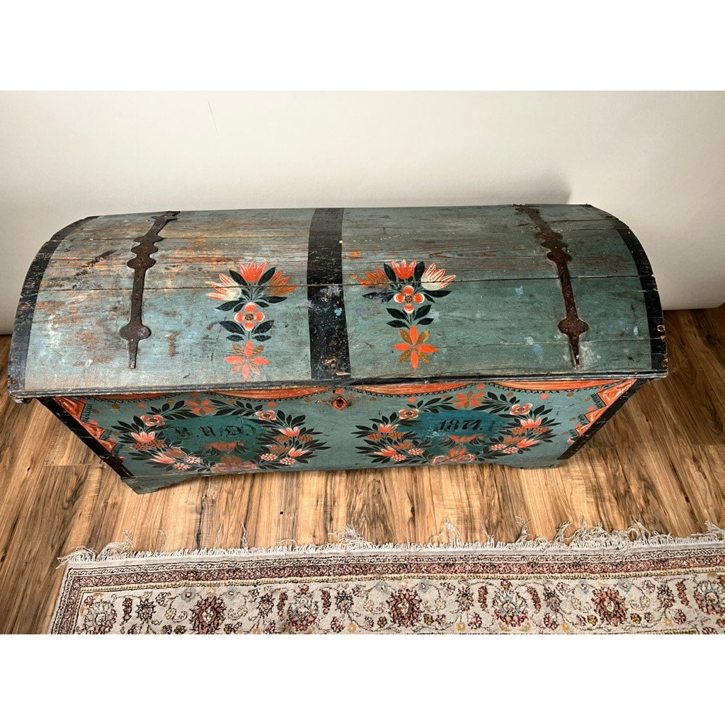 An Antique Swedish Oak Kista Dowry Chest c. 1841 with a curved lid and teal, red, and orange floral patterns rests on a wood floor beside a fringed, patterned rug. Brand: Unknown.