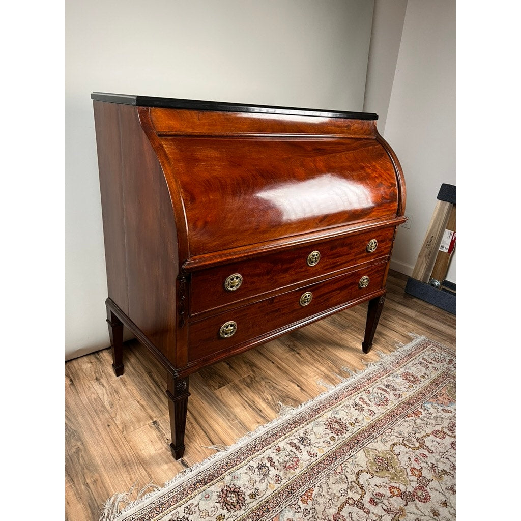 An Antique Directoire Style French Cylinder Desk with a slate top from the late 19th century stands on a wood floor beside a patterned rug and beige wall. The desk features elegant detailing and evokes classic French charm.