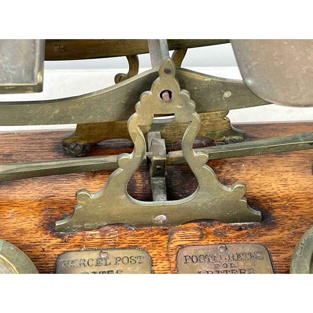 Close-up of an Antique 19th Century English Postal Scale with ornate brass details on a wooden base; metal tags labeled PARCEL POST and POSTAL RATES FOR LETTERS are partially visible at the bottom.