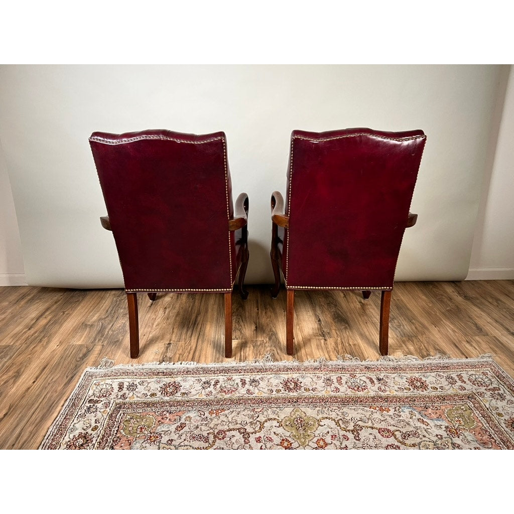 A pair of Chippendale Style Tufted Burgundy Leather Library Armchairs, featuring elegant wooden arms and legs, are positioned side by side with their backs to the camera on a wooden floor, complemented by a patterned area rug in front.