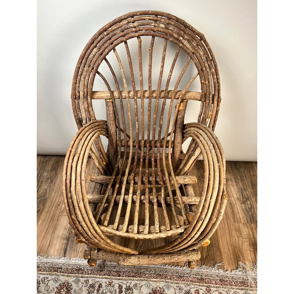 A Vintage Adirondack Bent Willow Arm Chair, crafted from curved and straight branches with a rounded back and armrests, is placed on a hardwood floor beside a patterned rug and a plain light-colored wall.