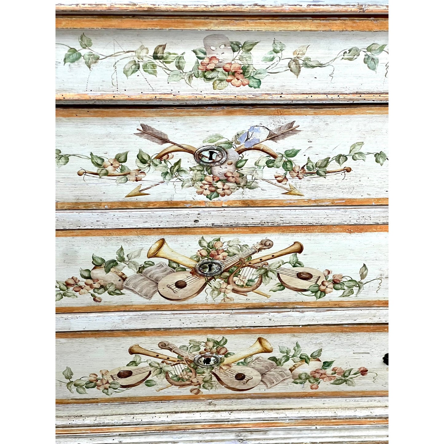 The Antique Italian Paint Decorated Four Drawer Commode c. Early 19th Century showcases hand-painted floral garlands and musical instruments in soft pastels on a distressed ivory background, reflecting classic Italian provincial style.