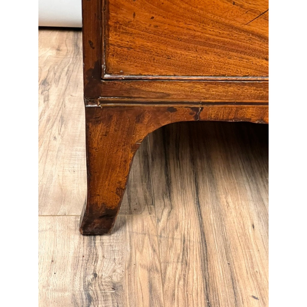 A close-up of the lower corner and French feet of an Antique George III Two Over Four Chest, c. early 19th century, highlighting its polished mahogany wood resting on a light-colored wood floor. Brand: Unknown.