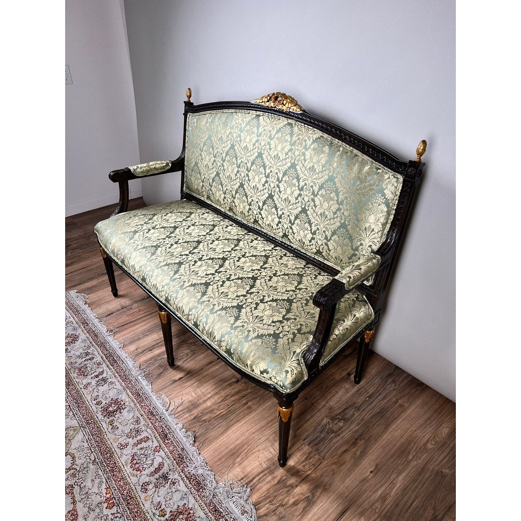 Vintage Louis XVI style settee in green damask upholstery, featuring ornate gold accents, sits on a wooden floor beside a patterned rug against a plain white wall. Brand: None/Unknown.