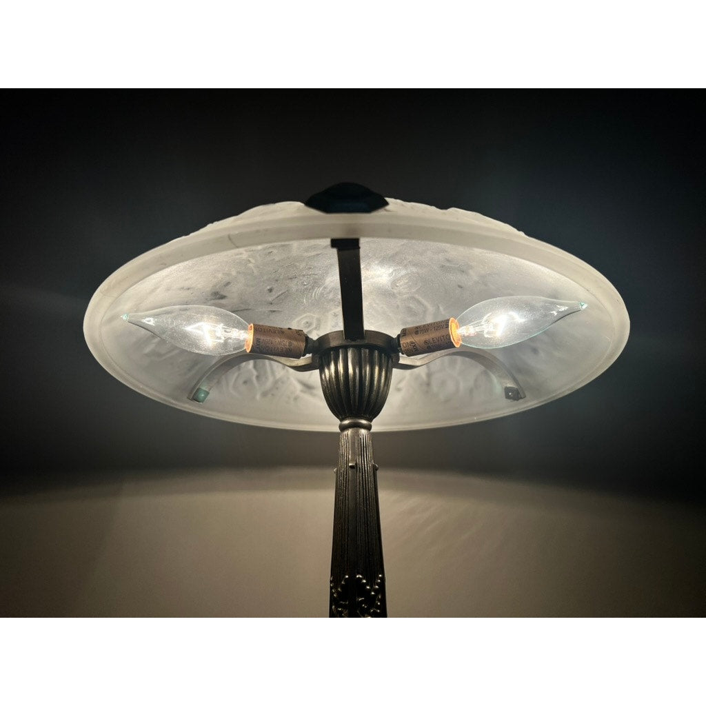 A close-up of a Vintage French Art Deco Frosted Glass Table Lamp by Muller Frères Lunéville c. 1920 displays three glowing bulbs beneath its frosted shade, illuminating a dark room with warm light.