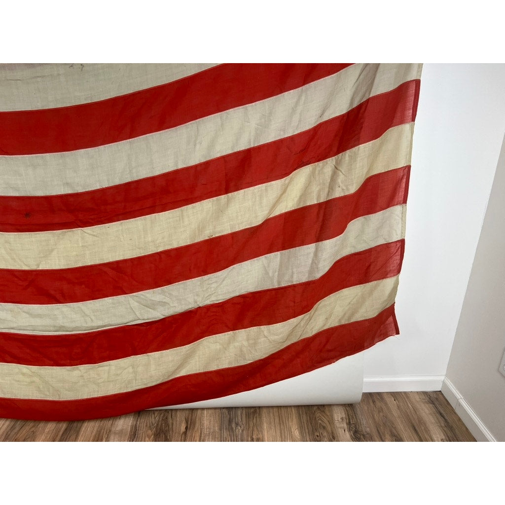 A close-up of an Antique 45 Star American Flag from the Spanish American War Era (9’9 x 6) featuring wide, horizontal red and off-white stripes, draped against a white wall and wood floor. Brand: None/Unknown.