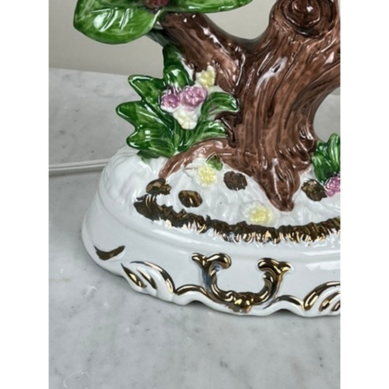 Close-up of the base of a Vintage Lena Liu Porcelain Hummingbird Accent Lamp, featuring a brown tree trunk, green leaves, pink and yellow flowers, and a white base with gold and brown accents on marble. Brand: Unknown.