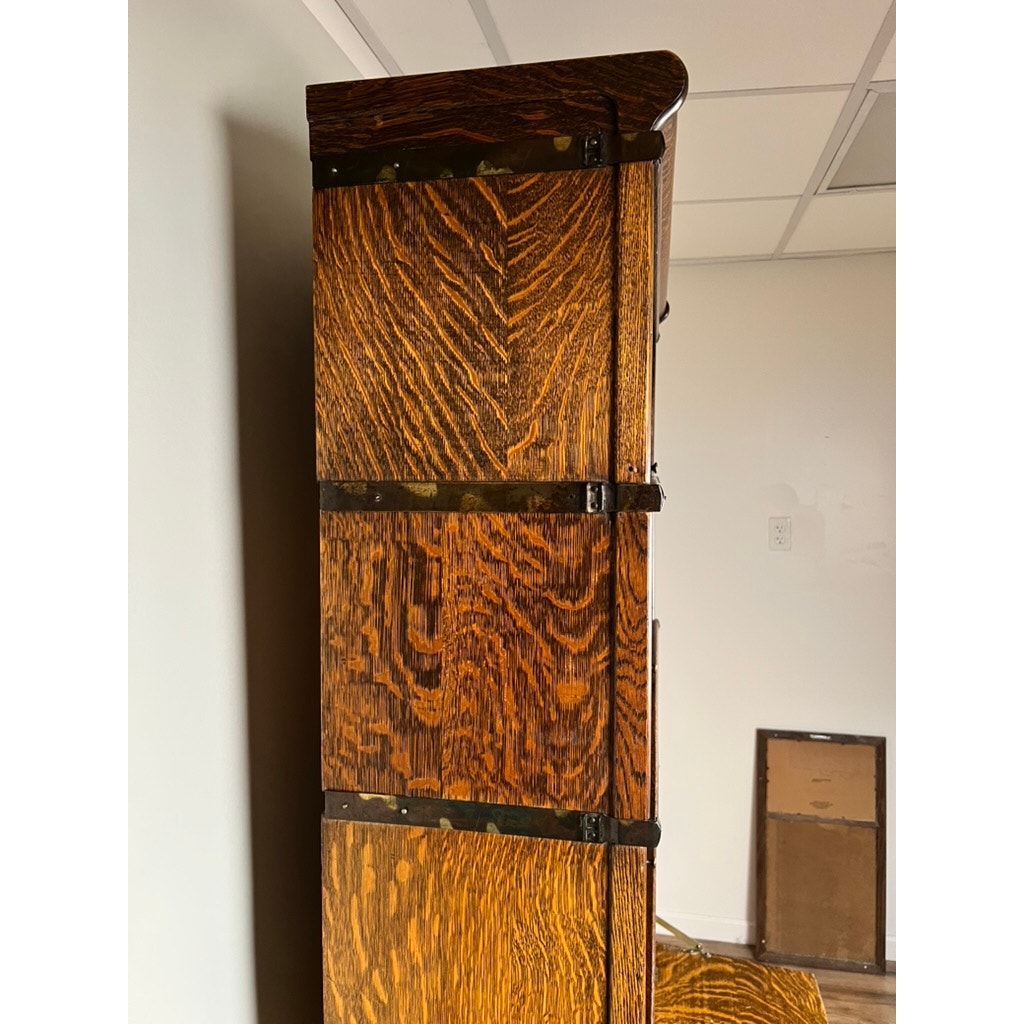 A close-up side view of the Antique Globe-Wernicke Quarter Sawn Oak 6-Stack Barrister Bookcase Secretary Desk (c. 1905–1915) by Globe Wernicke, showcasing its tall silhouette crafted from quarter-sawn oak with a rich, varied grain pattern and dark metal hinges, reminiscent of classic barrister bookcases, placed against a light-colored wall beneath a tiled ceiling.