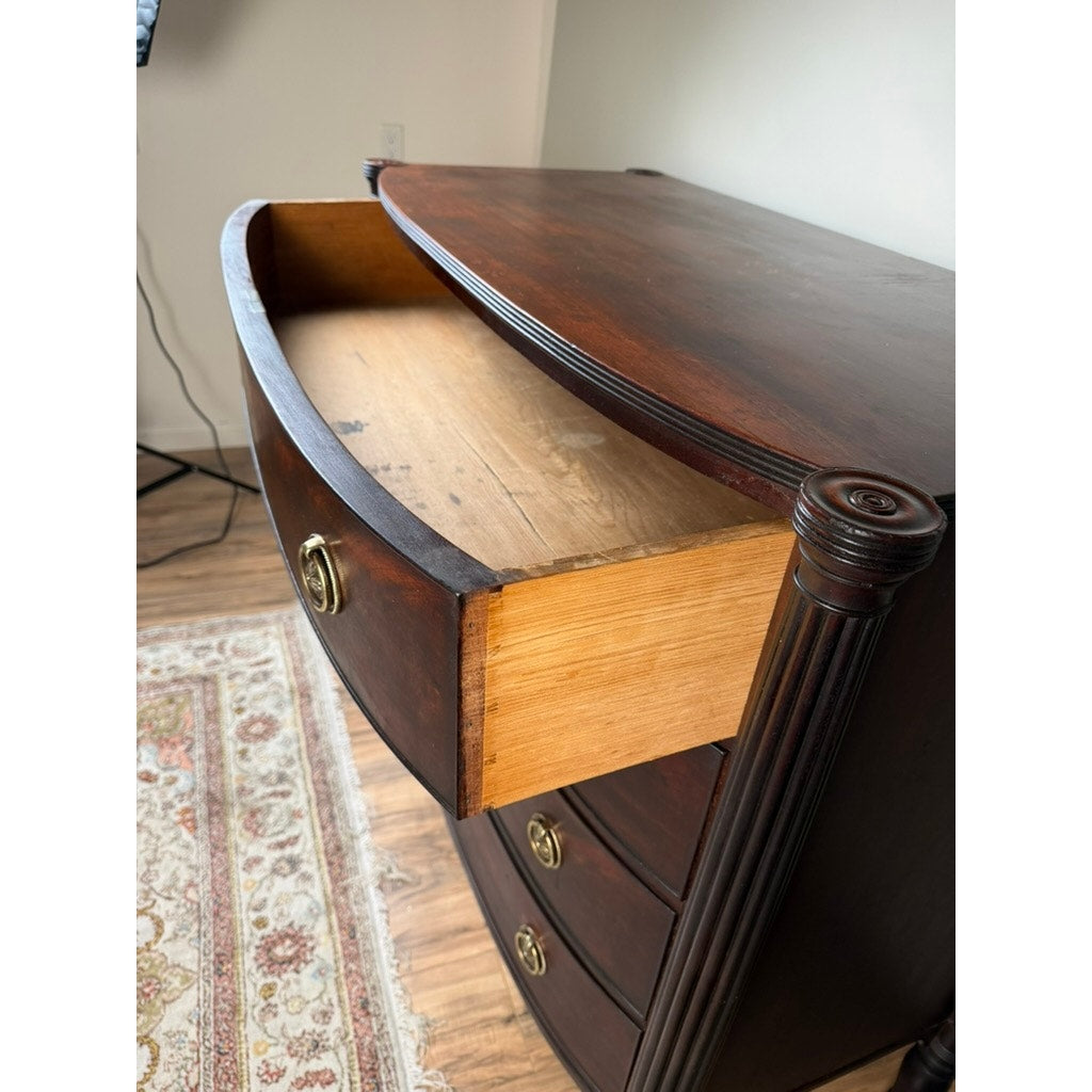 An Early 19th Century New England Federal Mahogany Bow-Front Chest of Drawers, c. 1810–1825, with its gracefully curved front, dark mahogany finish, and refined details evokes the elegance of Federal-era craftsmanship. The open top drawer reveals the chest’s interior, while a patterned rug can be seen on the floor beside it.