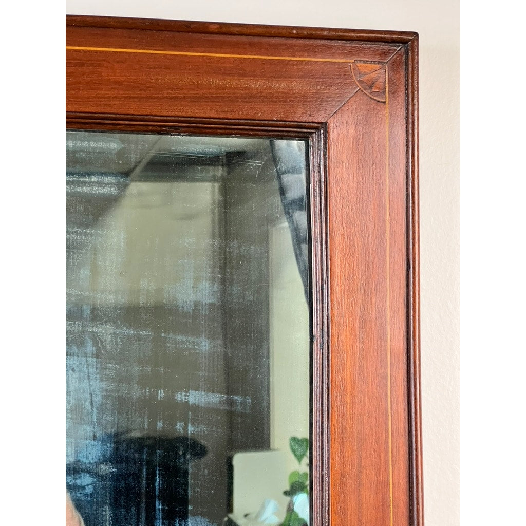 Close-up of the top right corner of an American Federal Mahogany Wall Mirror with fan inlay, featuring visible wood grain, a small crack, faint streaks on the original hand silvered glass (circa 1810–1825), and a blurred reflection of an indoor room with some plants.