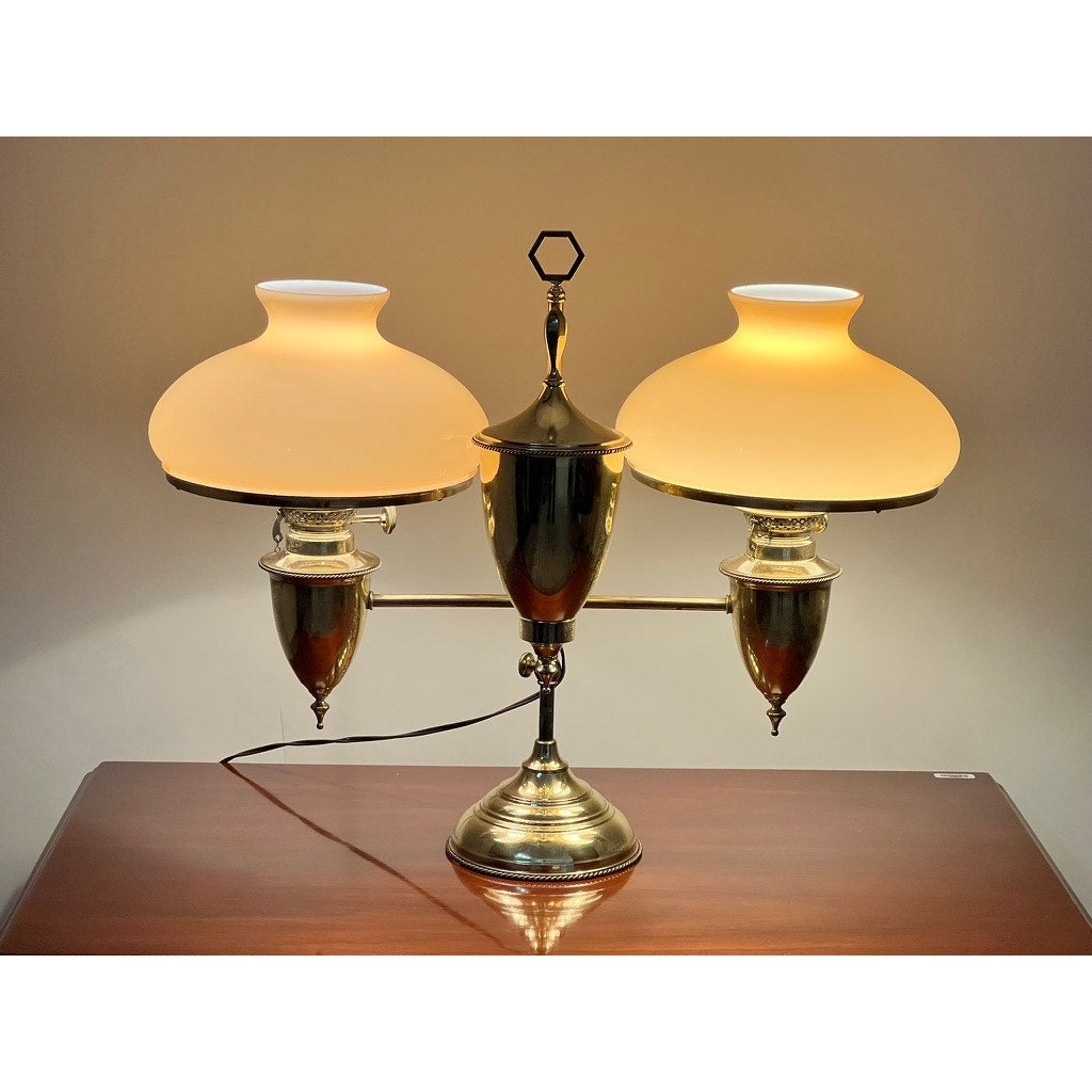 The Antique Bradley and Hubbard Brass Double-arm Student Lamp features two carmel shades glowing warmly on a polished wooden surface, capturing vintage charm from the Bradley and Hubbard brand against a softly lit background.