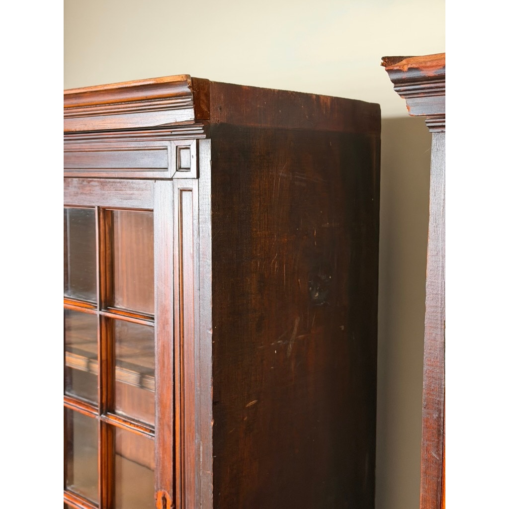 Close-up of the side of a Pair of Mid-19th Century New England Cherry Glazed Display Cupboards, c. 1840–1860 by Pearl City Antiques, highlighting rich cherry wood grain, visible scratches, and decorative trim near the top—distinctive details typical of early American design. Another matching cupboard from the pair is partially visible on the right.