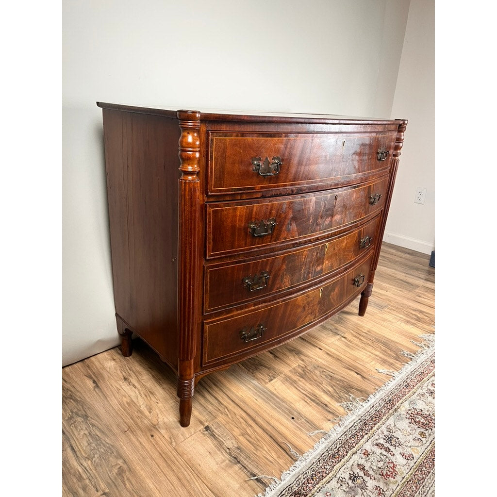 An Antique Sheraton Mahogany Bowfront Chest c. Early 19th Century with four curved drawers, ornate handles, and side columns stands on a wooden floor beside a patterned rug and light-colored wall. Brand: Unknown.