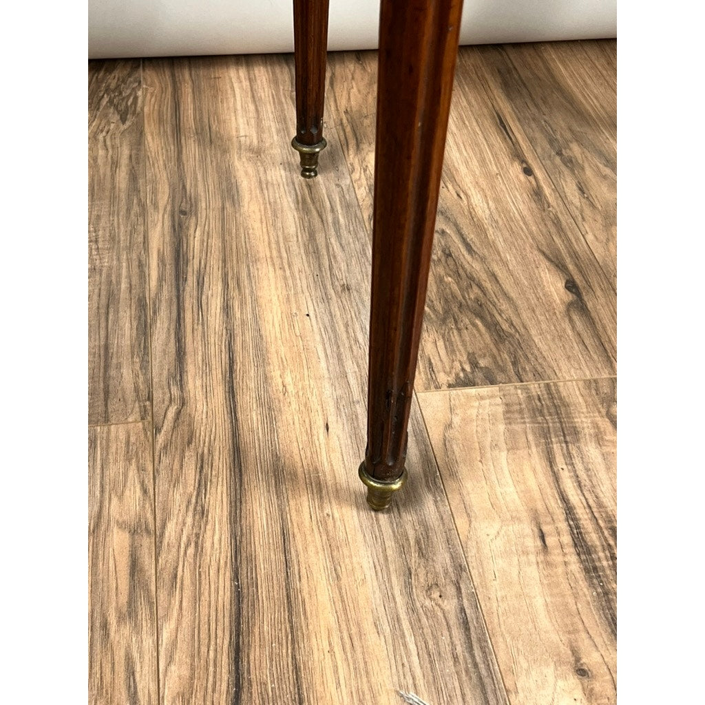 Close-up of two slender, tapered mahogany legs with brass caps from an antique 19th century French Louis XVI-style bouillotte table with marble top and pierced brass gallery, standing on a wood-patterned floor.