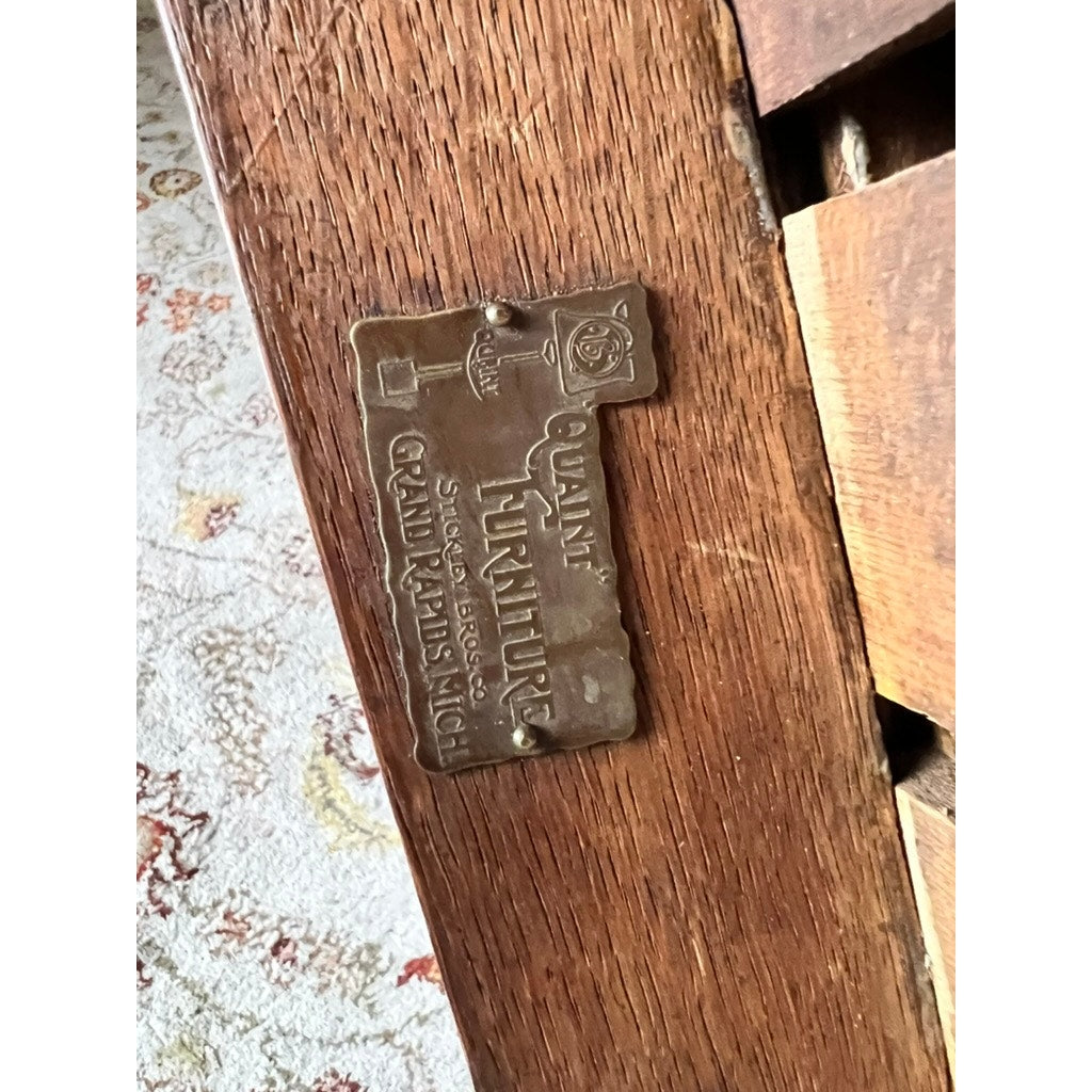 A brass plaque on this antique quarter sawn Mission oak arm chair by Quaint Furniture c. 1920 highlights the brand, with a patterned carpet seen behind the wooden piece.