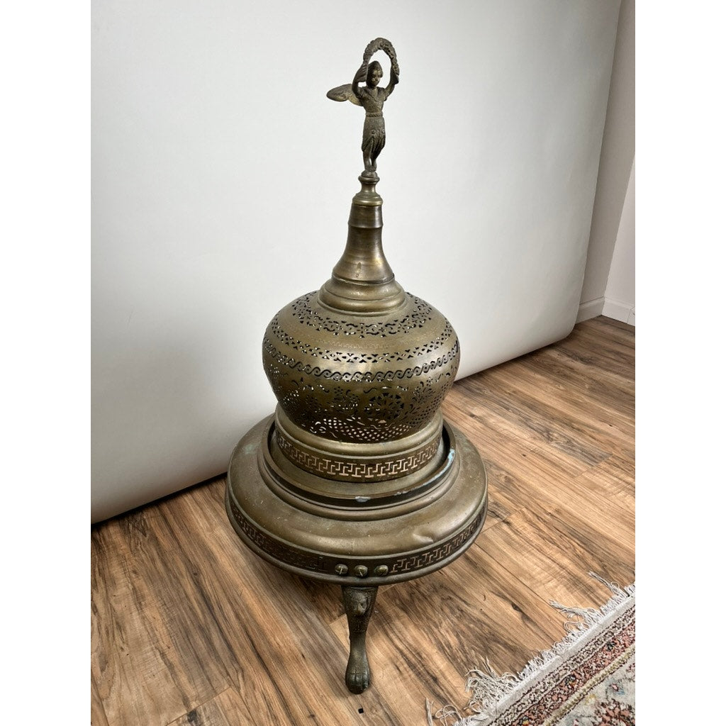 The Antique Venetian Brass Bell Room Heater Brazier (c. 19th Century) stands on three curved legs atop a wooden floor beside a white wall and patterned rug, functioning as both décor and an antique heater. Brand: Unknown.