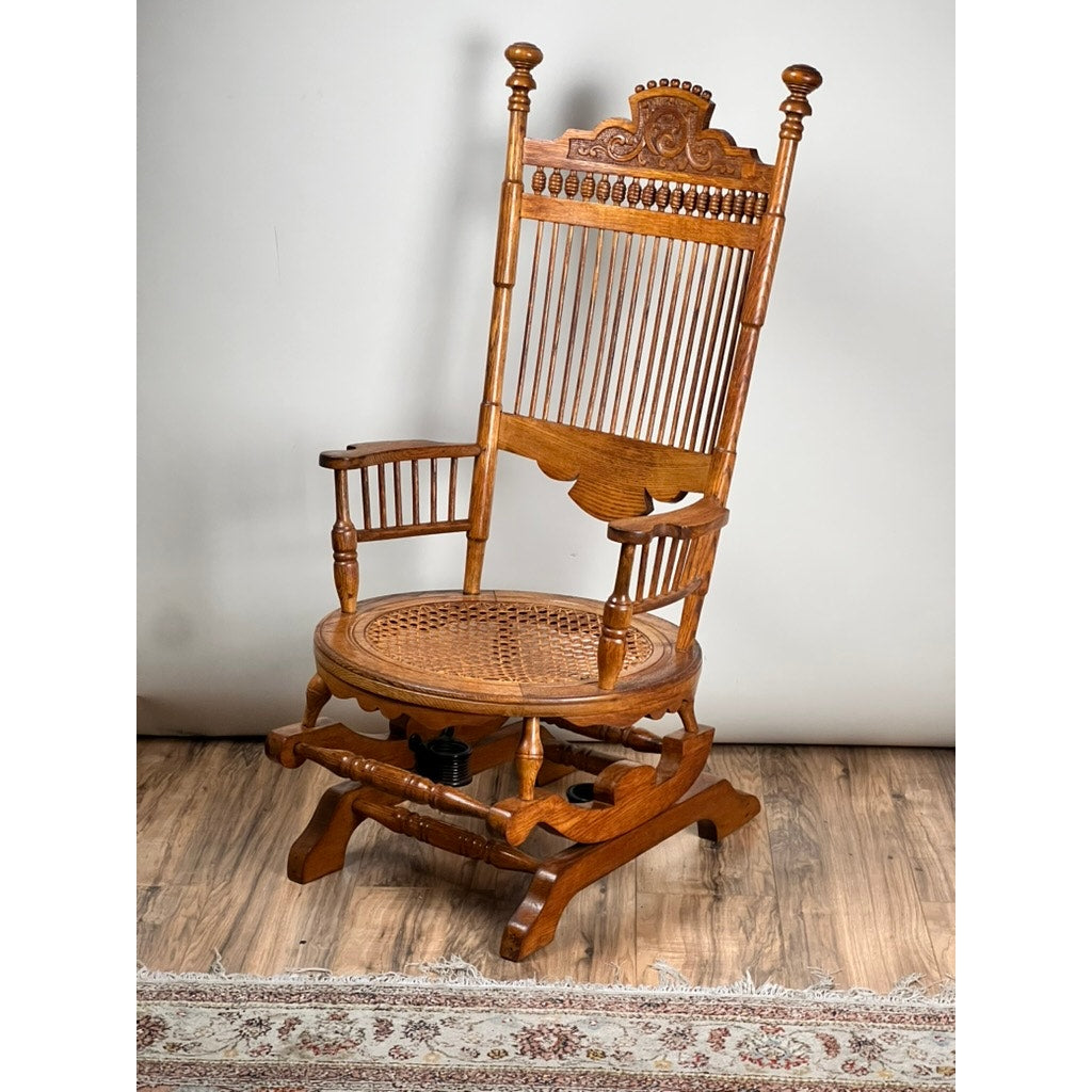 An Antique Victorian Oak Platform Rocker from the late 19th century, featuring ornate carvings and a spindle back, sits near a patterned rug against a plain light-colored wall—an elegant vintage accent.