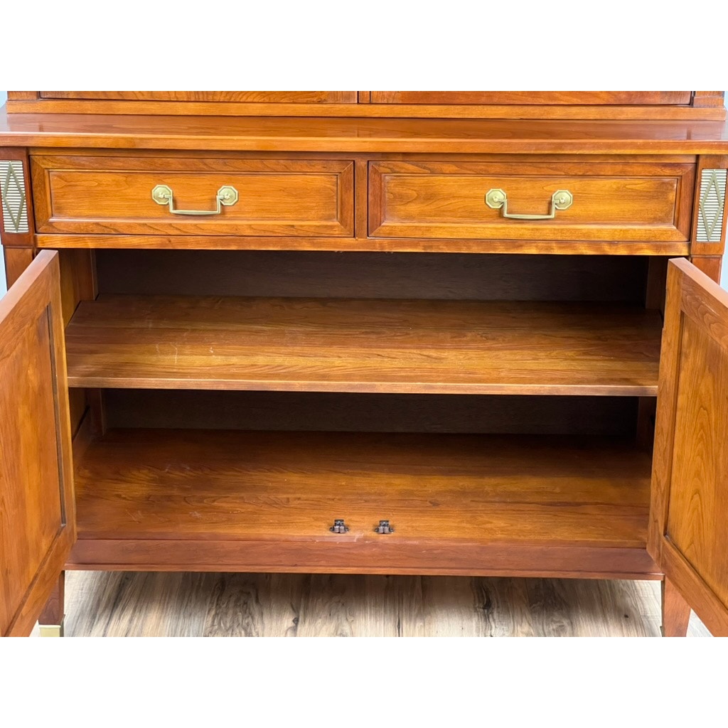 A Vintage William IV Style Cherry Glass Door Bookcase by Stickley with empty shelves and two drawers with brass handles stands on a wooden floor.