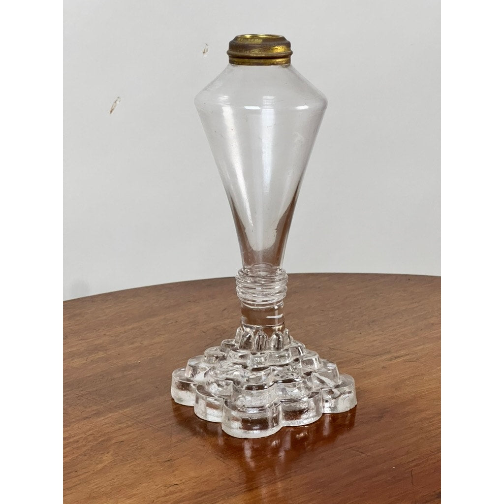 An Attributed Boston & Sandwich pressed glass boudoir fluid lamp, circa 1850, sits on a wooden surface. Reminiscent of mid-19th century design, it features a clear faceted body with a geometric, stepped square base and a brass burner—echoing the elegant style of Victorian pressed glass lamps. No wick or chimney is attached.
