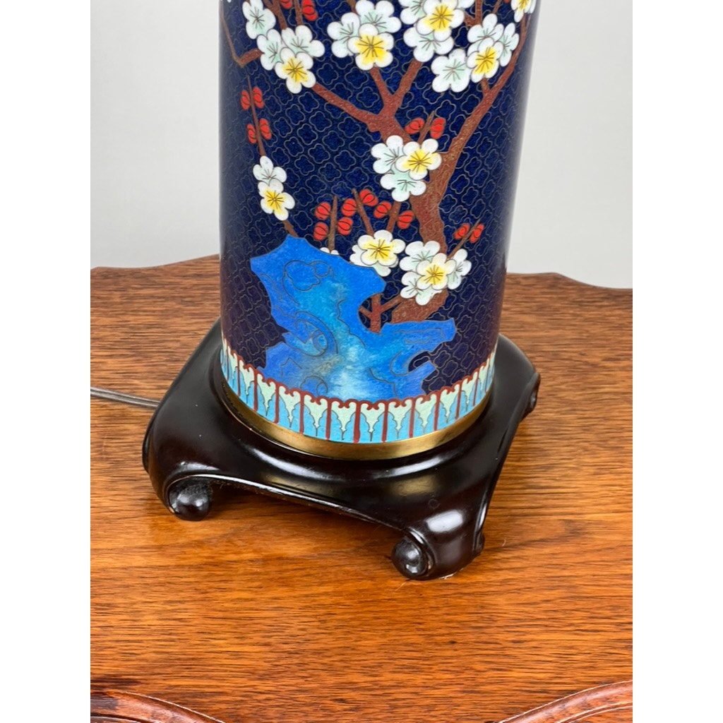 A close-up of the Vintage Japanese Cloisonné Cylinder Table Lamp by Knob Creek, featuring white and red floral designs on a blue background, displayed on a dark wooden stand atop a brown wood surface.